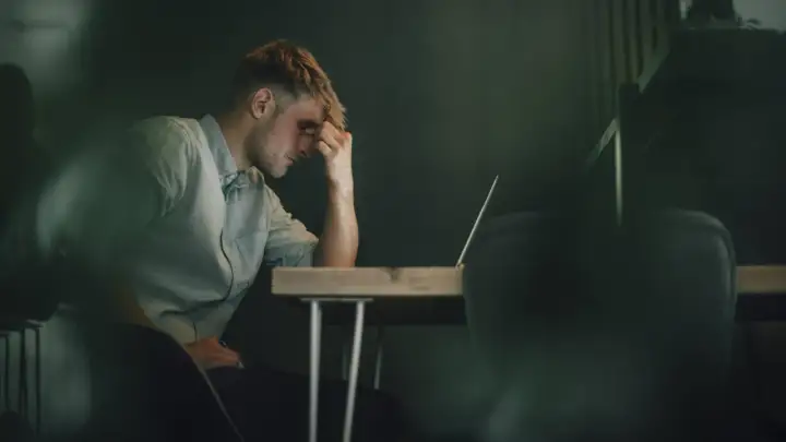 Ein Mann sitzt mit der Hand an der Stirn vor einem Laptop. Er wirkt nachdenklich, aber es ist möglich, dass er trotz Depressionen arbeitet.