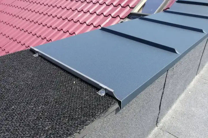 Szary ogniomur z blachy chroni dach przed ogniem, sąsiadując z czerwonym dachem i czarną papą.