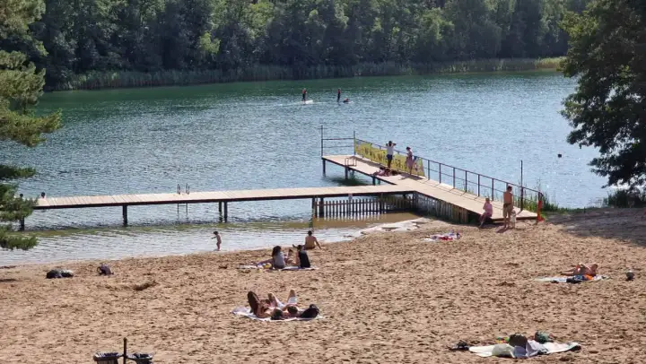 Słoneczna borówno plaża nad jeziorem, ludzie odpoczywają na piasku i w wodzie, niektórzy pływają na desce SUP.