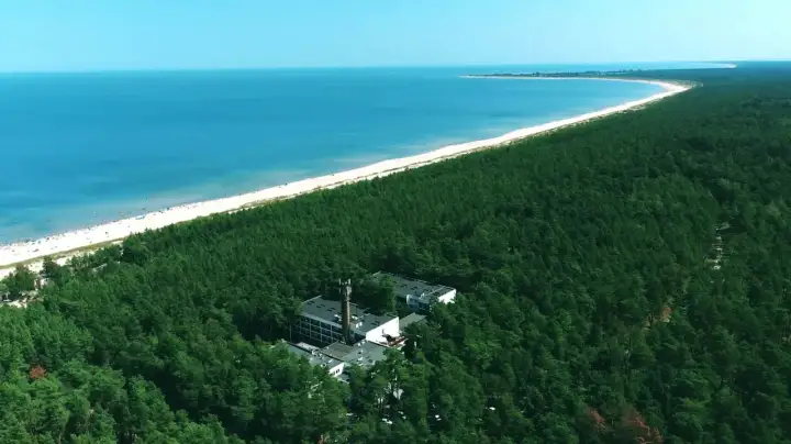 Widok z lotu ptaka na hotel otoczony lasem sosnowym, z plażą i morzem w tle.