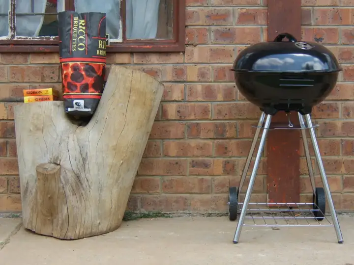 Grill stoi obok pnia, a torba z węglem drzewnym wisi na pniu. Odległość grilla od granicy działki jest bezpieczna.