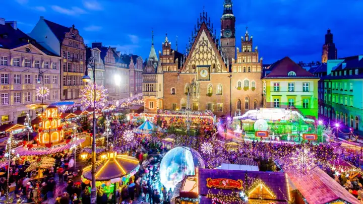 Tłoczny jarmark we Wrocławiu nocą, rozświetlony tysiącami lampek. Widać Ratusz, karuzelę i stragany.