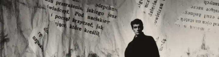 Młody mężczyzna, być może Hanuszkiewicz, stoi na tle podartych kartek z tekstem.