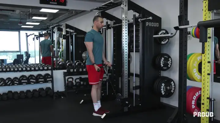 Mężczyzna wykonuje ćwiczenia na biceps przy użyciu wyciągu. W tle widoczne są sztangi, hantle i talerze obciążeniowe.