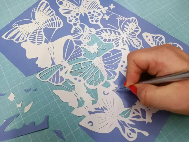 Main d'une personne aux ongles rouges découpant avec précision un motif de papillons en papier. Un tuto de papier découpé en cours.