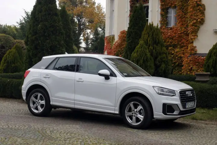 Białe Audi Q2 na brukowanym podjeździe, otoczone zielenią i jesiennymi liśćmi. Idealne do miasta, jak sugerują pierwsze audi q2 opinie.