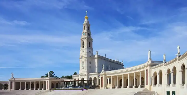 Biała bazylika z wieżą i złotą koroną na szczycie, otoczona kolumnadą. To Sanktuarium Matki Bożej Różańcowej w Fatimie, miejsce pielgrzymek.
