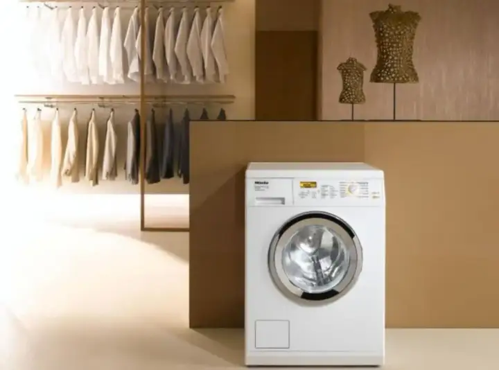 Lavatrice Miele bianca con ingresso acqua calda e fredda, accanto a un armadio con abiti ordinati.
