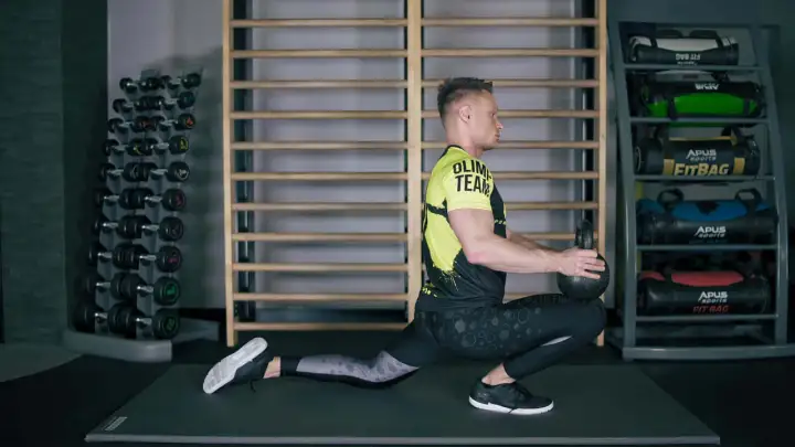 Mężczyzna wykonuje ćwiczenia mobilizacyjne z kettlebell, pogłębiając wykrok. W tle stojaki z hantlami i fitbagami.