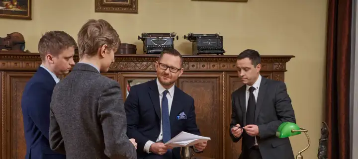 Czterech mężczyzn w garniturach w gabinecie, dyskutujących nad dokumentami.