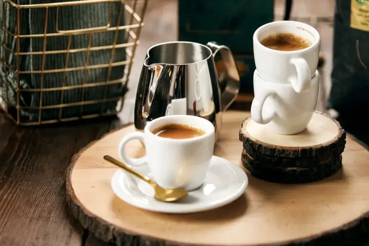 Två koppar espresso, en med en guldig sked, och en mjölkkanna på ett träfat. Perfekt för en stunds njutning med espresso bryggkaffe.
