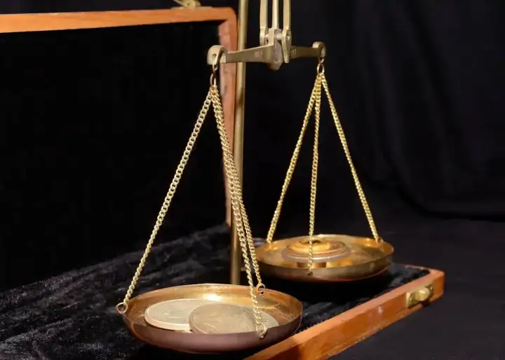 Waga szalkowa z monetami i ciężarkami. Na jednej szalce leżą monety, na drugiej ciężarki, symbolizujące uncję złota.