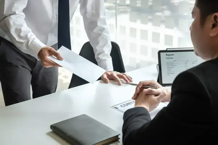 Przełożony wręcza pracownikowi dokumenty, być może umowa na czas określony. Na stole leży teczka i tablet z raportem.