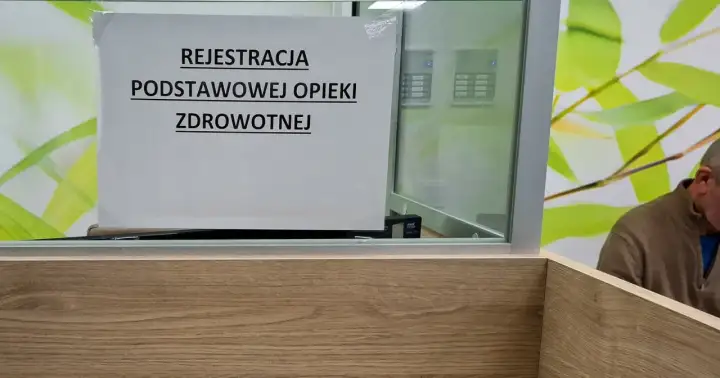 Tabliczka "Rejestracja podstawowej opieki zdrowotnej" w poczekalni. W tle widać fragment mężczyzny i zielone liście.