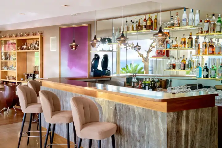 Un **majestic bar** avec des tabourets beiges, des étagères remplies de bouteilles et une vue sur la mer.