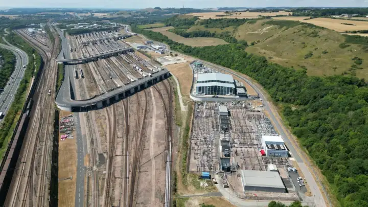 Rozległa infrastruktura kolejowa i stacja transformatorowa przy Eurotunelu. W tle wzgórza z białym koniem.