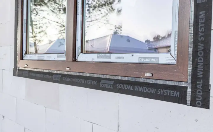 Brązowe okno w trakcie ciepłego montażu z użyciem taśmy SOUDAL WINDOW SYSTEM. Widoczna biała ściana z bloczków.