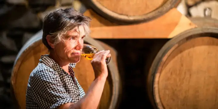 Une femme déguste un verre de vin de Pau, entourée de fûts de chêne dans une cave.