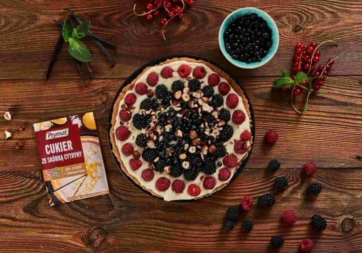Pyszna tarta z mascarpone, malinami, jeżynami i borówkami, posypana orzechami. Obok opakowanie cukru z skórką cytrynową.