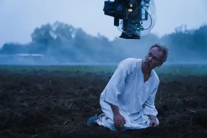 Syn Macieja Boryny klęczy na zaoranym polu. Nad nim wisi kamera filmowa.
