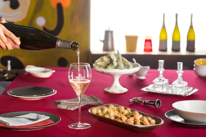 Versando vino tipico della Franciacorta in un calice, su un tavolo apparecchiato con stuzzichini e bottiglie.