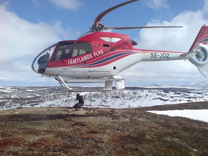 Czerwony helikopter Jämtlands Flyg SE-JOZ wylądował na zaśnieżonym wzgórzu. Fotograf robi zdjęcie, uchwycony przez efekt rolling shutter.