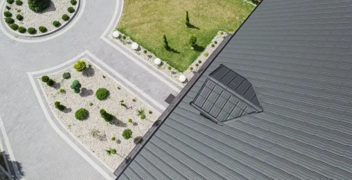 Nowoczesny dach pulpitowy z szarej blachy, z widocznym oknem dachowym. Obok zadbany ogród z brukowanymi ścieżkami i niską roślinnością.