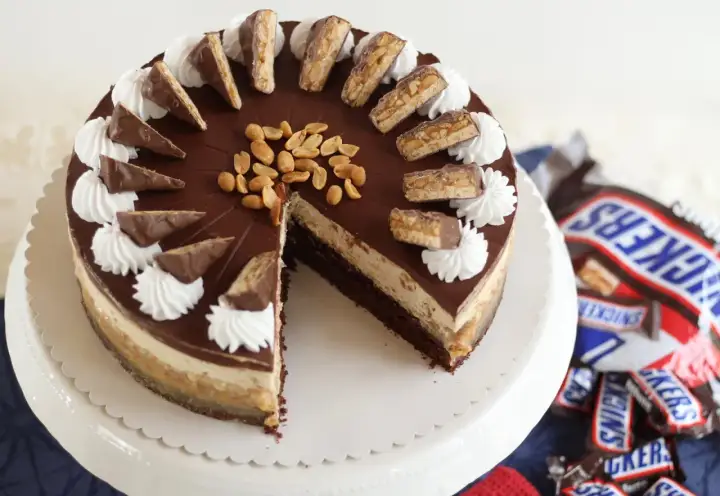 Eine köstliche Snickers Torte mit Schokoladenglasur, Erdnüssen und Sahnetuffs. Kleine Snickers-Stücke zieren den Rand.