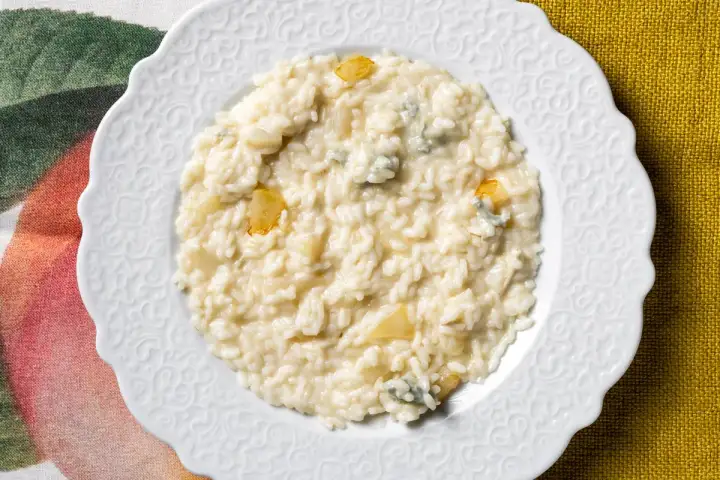 Un cremoso risotto pere e gorgonzola, servito su un piatto bianco decorato, con pezzetti di pera e formaggio blu visibili.