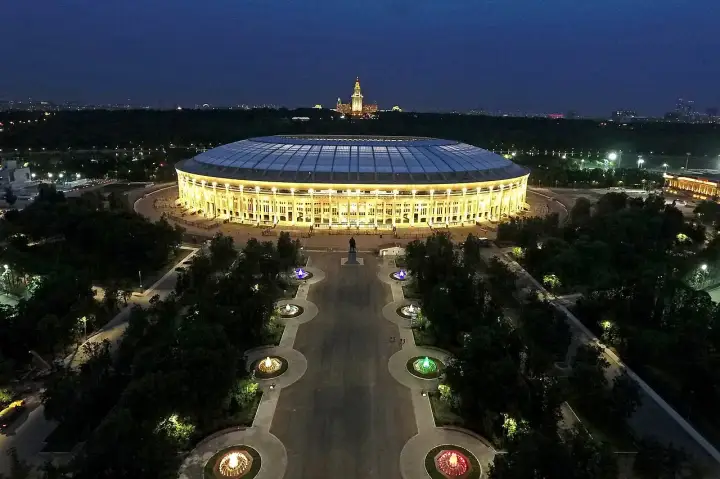 Oświetlony moskiewski stadion nocą, z aleją fontann i pomnikiem w tle.