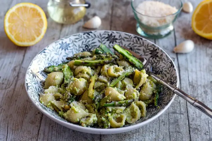 Un plat de pâtes aux asperges, garni de parmesan râpé, avec des citrons et de l'ail en arrière-plan.