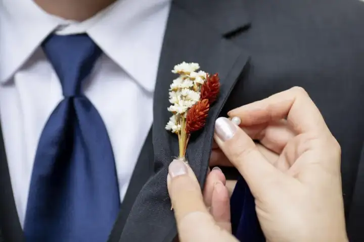 Dłoń przypina do marynarki mały bukiecik z suszonych kwiatów. To właśnie jest butonierka co to za ozdoba.