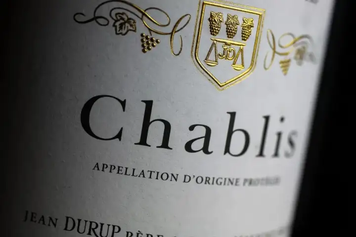Gros plan sur une étiquette de vin Chablis, avec un blason doré orné de grappes de raisin et d'une balance. L'appellation d'origine protégée est clairement visible.