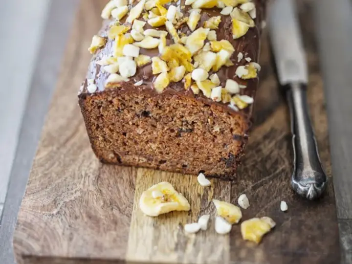 Saftiger bananenkuchen vegan mit Schokoladenglasur und Nüssen, auf einem Holzbrett mit einem Messer.