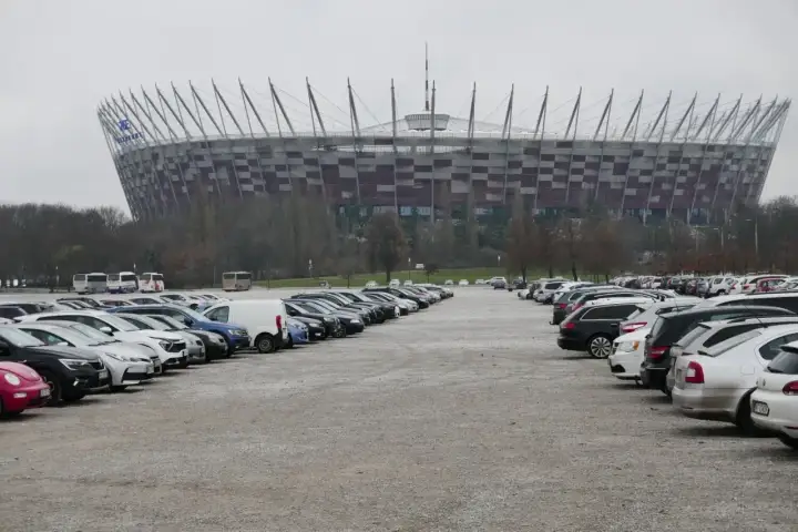Duży parking przed Stadionem Narodowym w Warszawie. Widać rzędy samochodów i autobusów na błoniach stadionu.