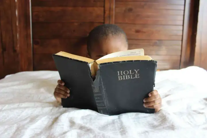 Dziecko z zaciekawieniem czyta Biblię, leżąc w łóżku. Słowa "Holy Bible" są widoczne na okładce.