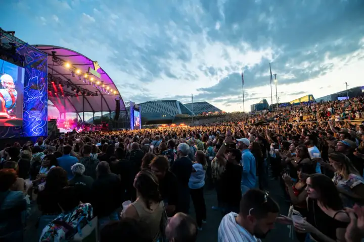 Multitud disfrutando de un concierto al aire libre. Se ve un gran escenario con luces y pantallas, y miles de personas animando. ¡Así se vive latino Zaragoza!