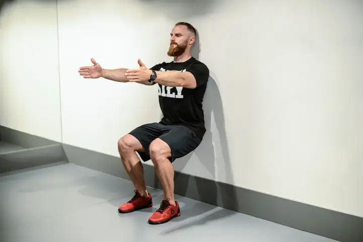 Mężczyzna z brodą wykonuje ćwiczenie wall sit, opierając plecy o ścianę i uginając nogi w kolanach.