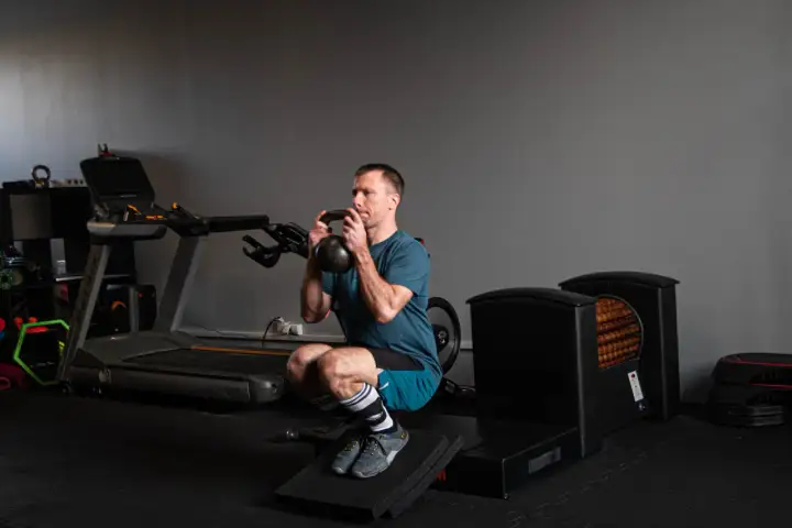 Kolarz wykonuje głęboki przysiad z kettlebell. W tle bieżnia i sprzęt do ćwiczeń.