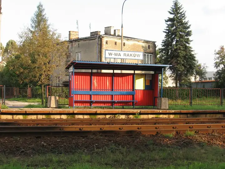 Przystanek kolejowy Warszawa Raków. Czerwona wiata z niebieską ławką i tablicą informacyjną. W tle budynek i wysokie drzewa.