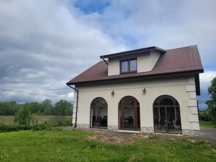 Nowy dom w Charsznicy z łukowymi oknami i brązowym dachem, otoczony zielenią i chmurami na niebie.