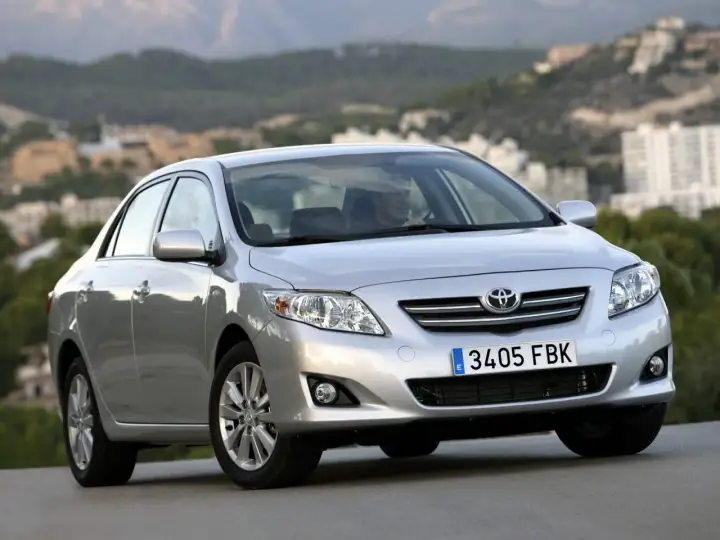 Srebrna Toyota Corolla 2010 z widocznym kierowcą, zaparkowana na tle krajobrazu z budynkami.