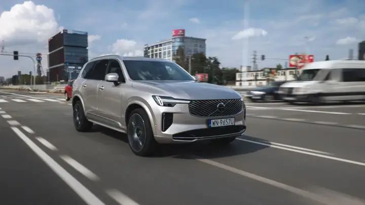 Srebrny Volvo XC90 T8 w ruchu miejskim, z nowoczesnymi budynkami w tle.