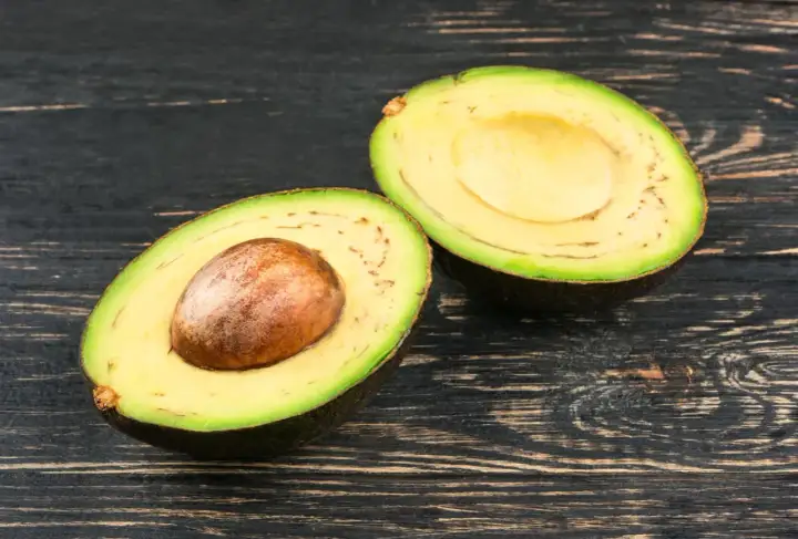 Eine aufgeschnittene Avocado mit Kern und einigen kleinen schwarzen Punkten im Fruchtfleisch liegt auf dunklem Holz.