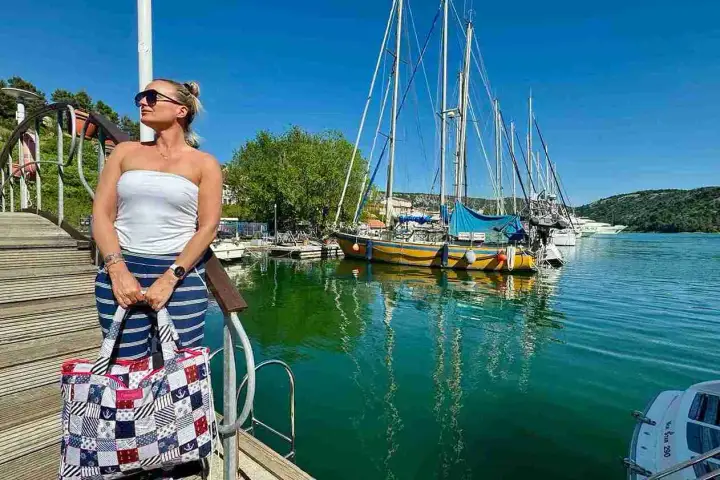 Kobieta z torbą w marynistycznym stylu na pomoście, w tle żaglówki i turkusowa woda. Idealny dzień na Chorwacji w maju.