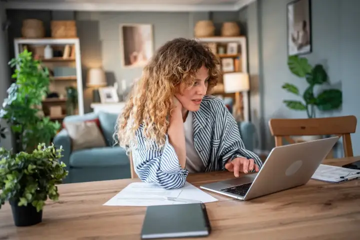 Donna con capelli ricci lavora al computer, documenti sul tavolo. Forse sta cercando informazioni su un mutuo con garante genitore per realizzare un sogno.