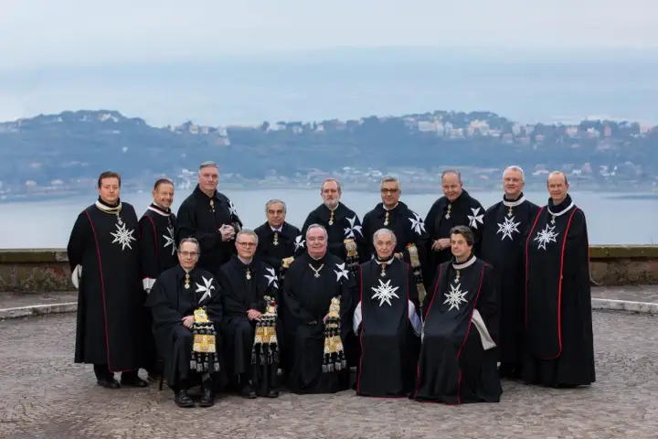 Uroczyste zgromadzenie członków zakonu maltańskiego w czarnych płaszczach z białymi krzyżami, na tle jeziora i miasta.