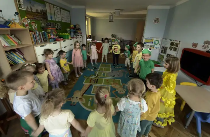 Dzieci trzymają się za ręce w kręgu na dywanie z zabawnymi zwierzątkami. To radosny dzień w **zespole szkolno przedszkolnym**.