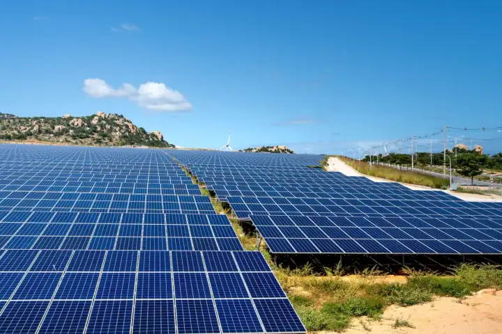 Extenso parque solar con paneles azules bajo un cielo despejado. En el fondo, colinas rocosas y una turbina eólica.