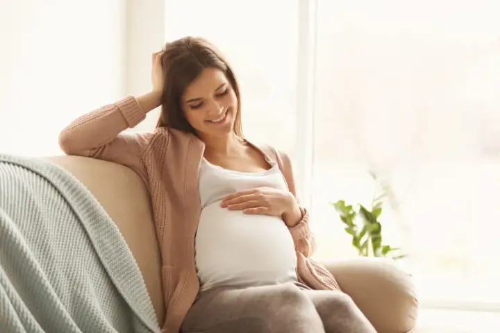 Uśmiechnięta ciężarna kobieta siedzi na kanapie, delikatnie dotykając brzucha. Myśli o bezpiecznym oczyszczaniu wodorowym w ciąży.